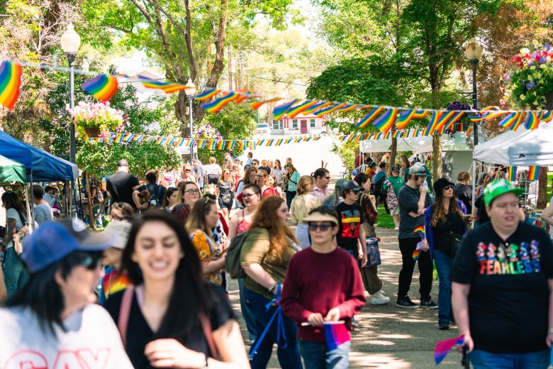 People, many of who are in colorful garb, walk down a path between booths and below lines of pride flags